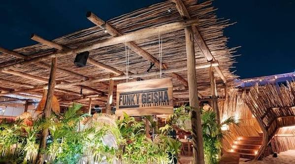 Outdoor tiki-style bar with a thatched roof, surrounded by lush green plants, and a hanging sign that reads 'FUNKY GEISHA' at night.