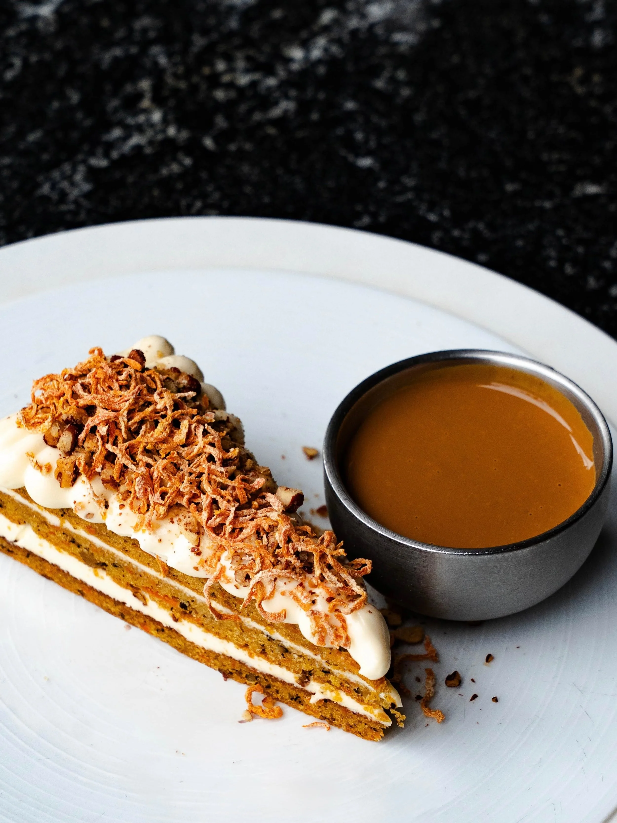 Slice of pumpkin pie topped with whipped cream and crushed toasted coconut, served with a small cup of caramel sauce on a white plate.