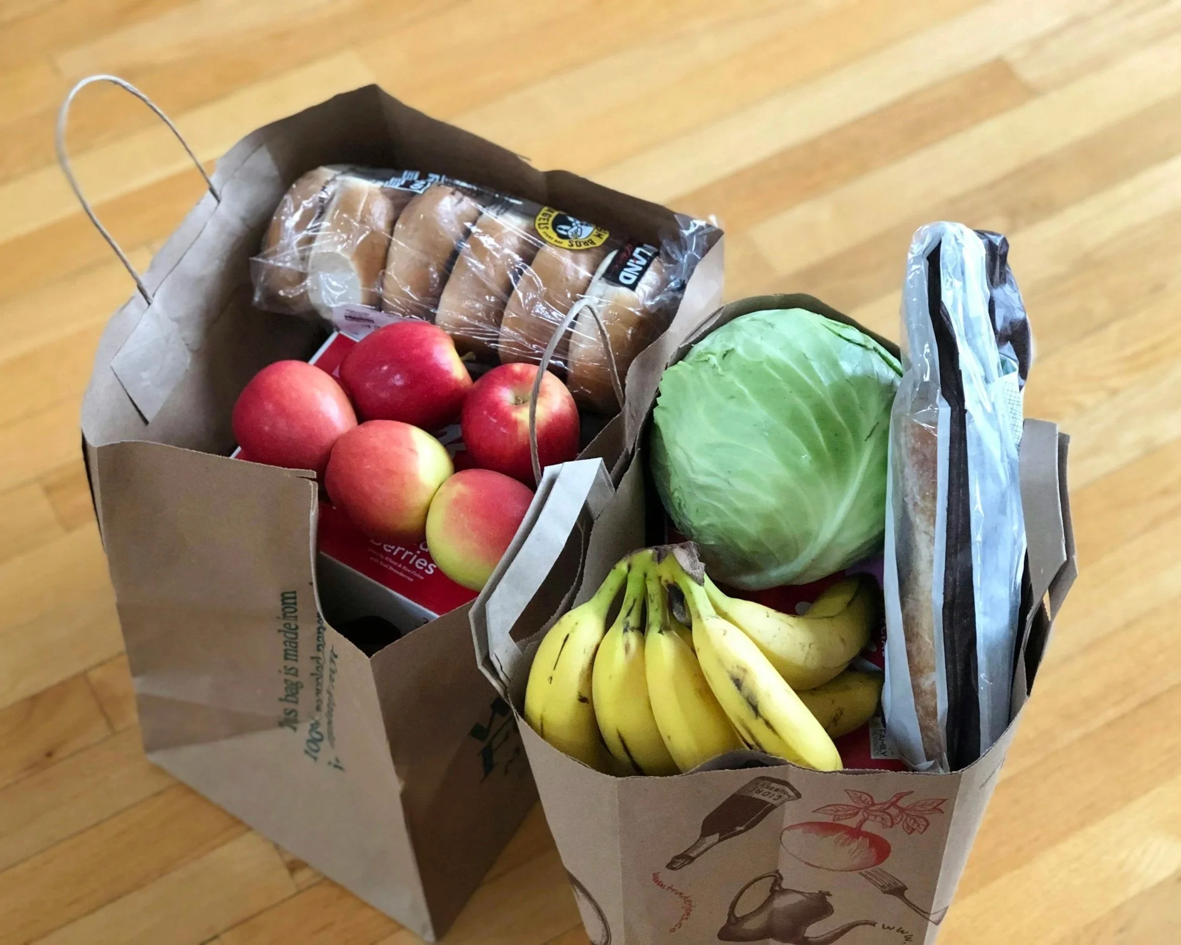 A paper grocery bag filled with red apples, bananas, a head of lettuce, a bag of sandwich bread, and a snack bag, on a wooden floor.