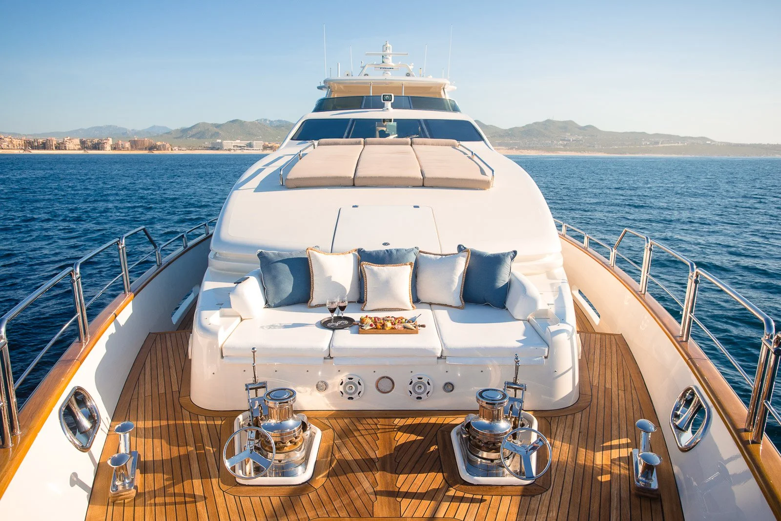 View of a luxurious yacht's deck with a seating area decorated with pillows, a tray of wine glasses and snacks, and scenic ocean and coastline in the background.