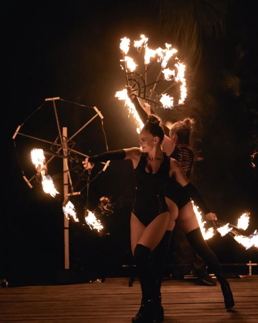 Two female fire performers in black costumes performing with flaming props on a wooden stage at night.