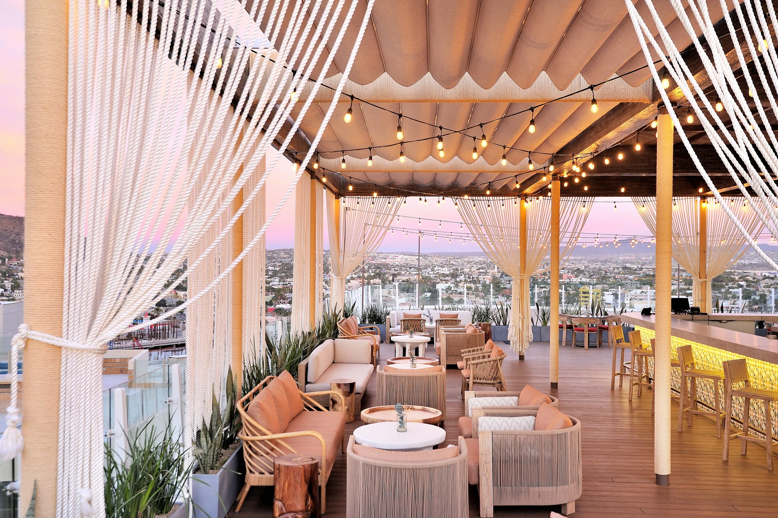Outdoor rooftop lounge with string lights, curtains, and city skyline view at sunset.
