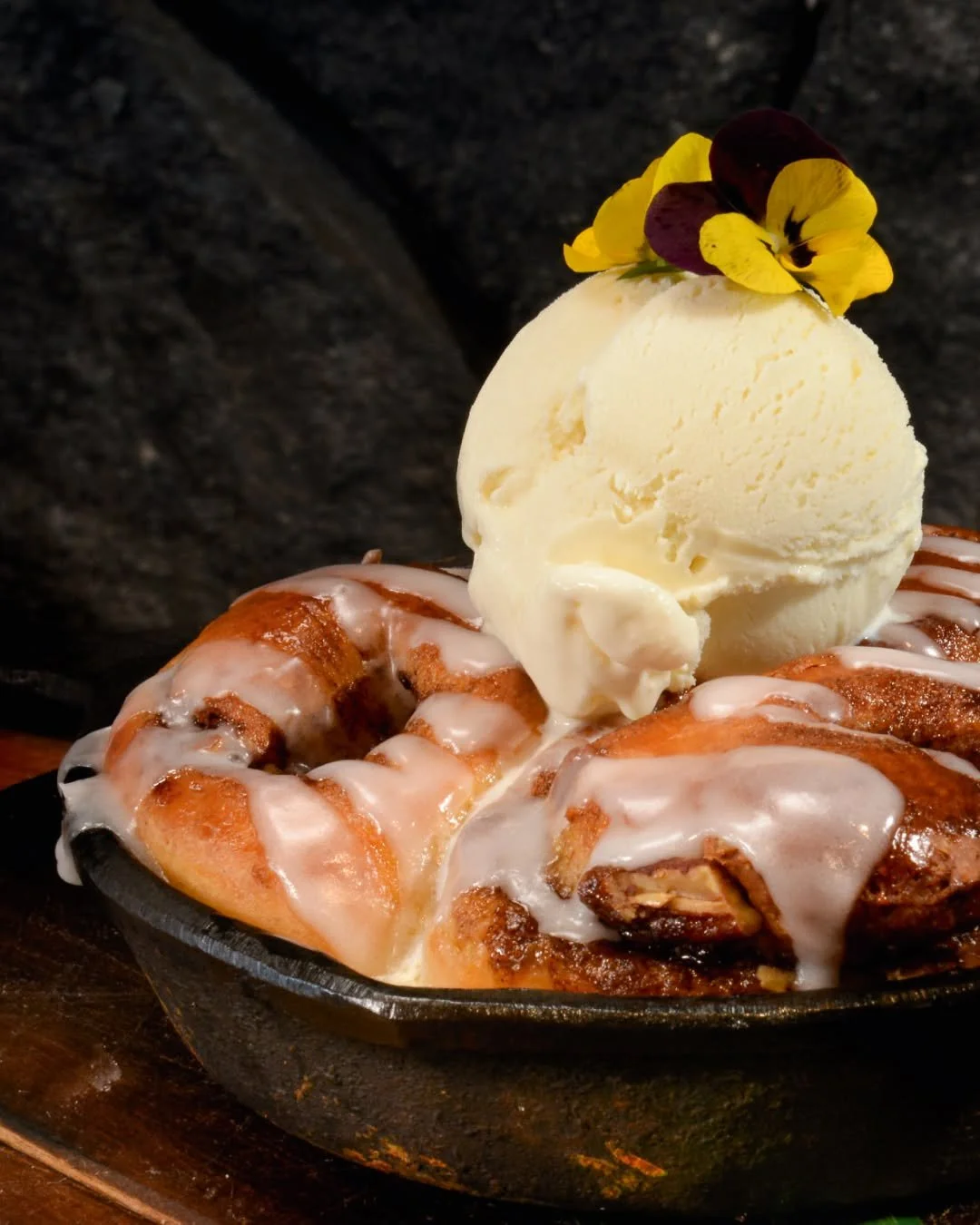Cinnamon roll topped with vanilla glaze, a scoop of vanilla ice cream, and edible flowers, served in a black cast iron skillet.