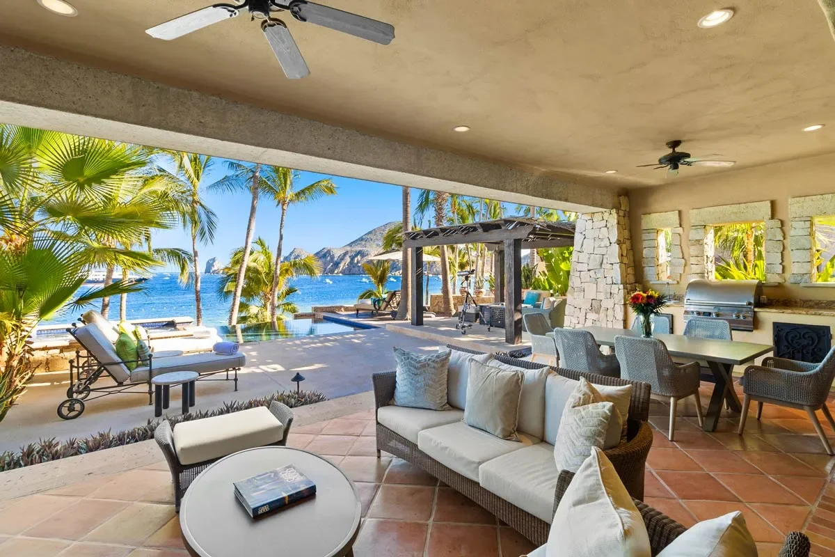 Luxury outdoor living space with seating area, dining table with flowers, kitchenette, ceiling fans, overlooking a tropical beach with palm trees, ocean, and mountains.