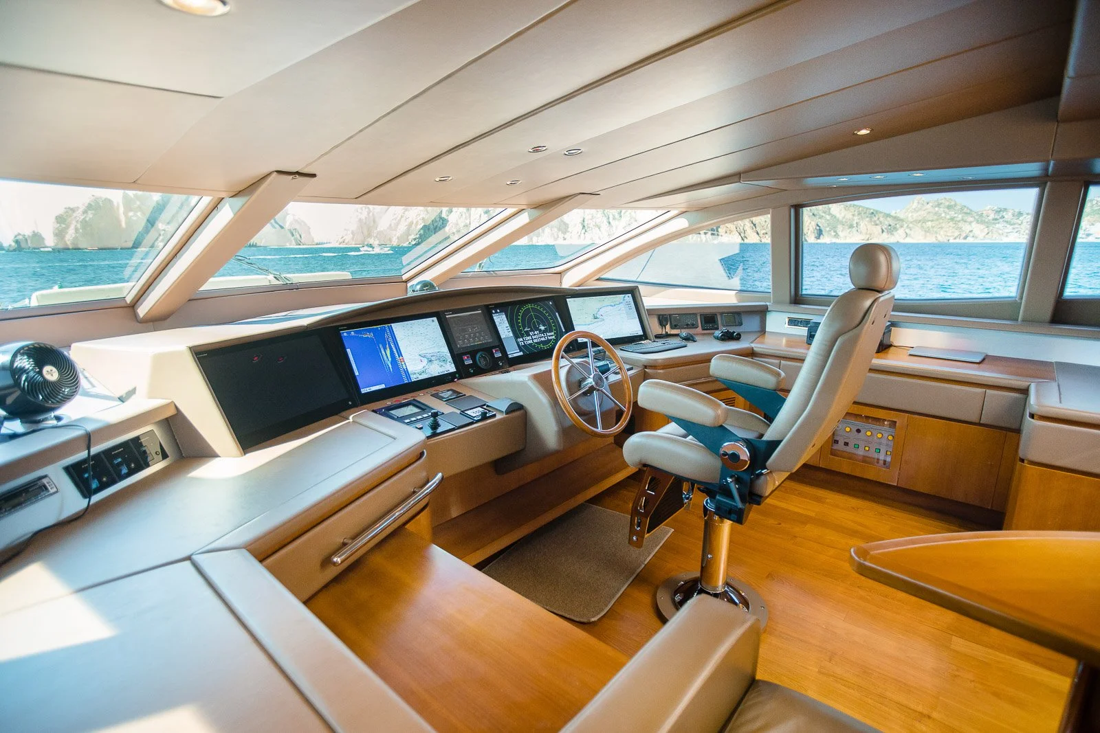 Interior view of a yacht steering room with control panels, a chair, and large windows showing a sea and coastline.