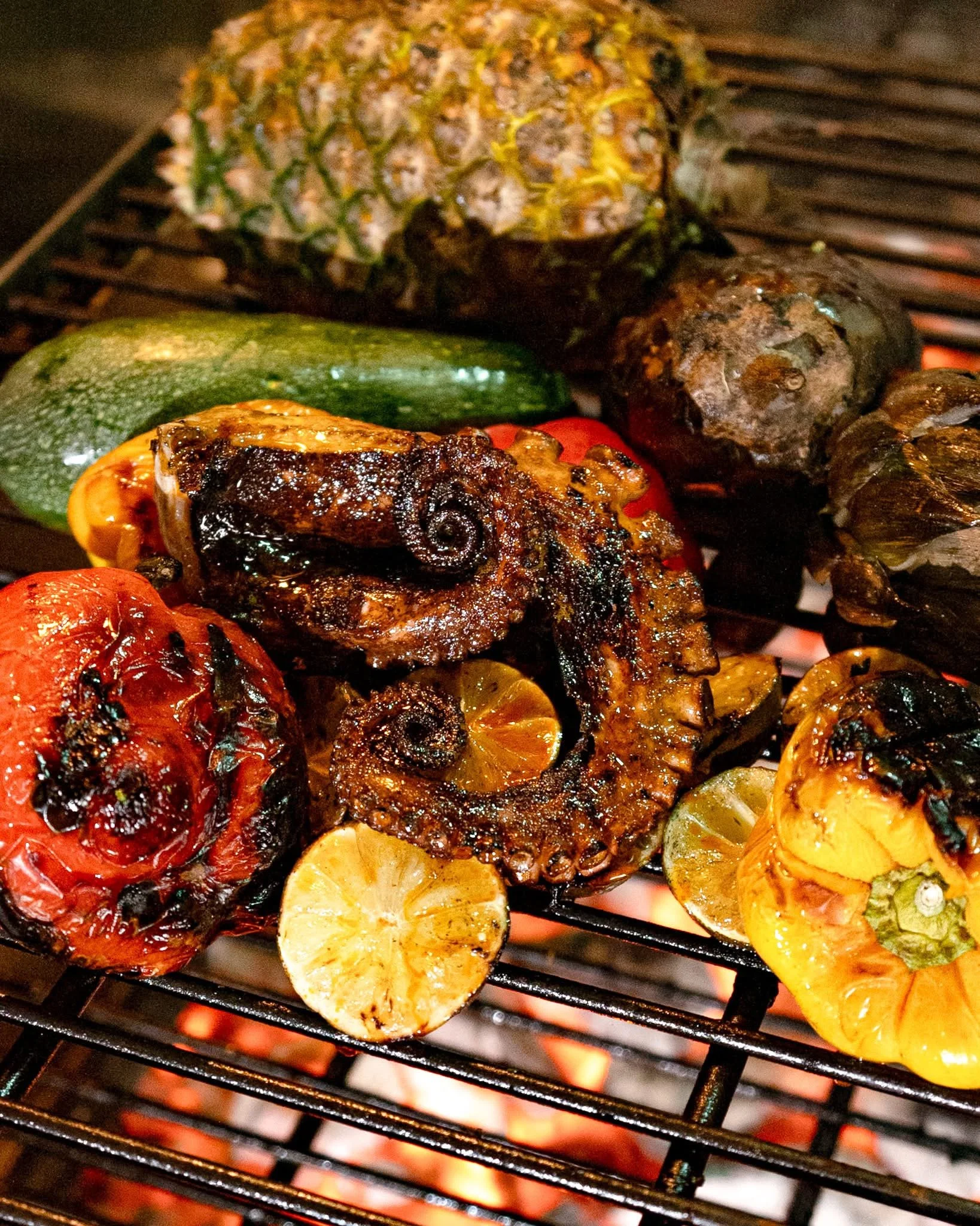 Close-up of vegetables and meat skewers grilling on a barbecue grill over open flames, including pineapple, zucchini, cherry tomatoes, mushrooms, lemon slices, and chunks of beef or lamb.
