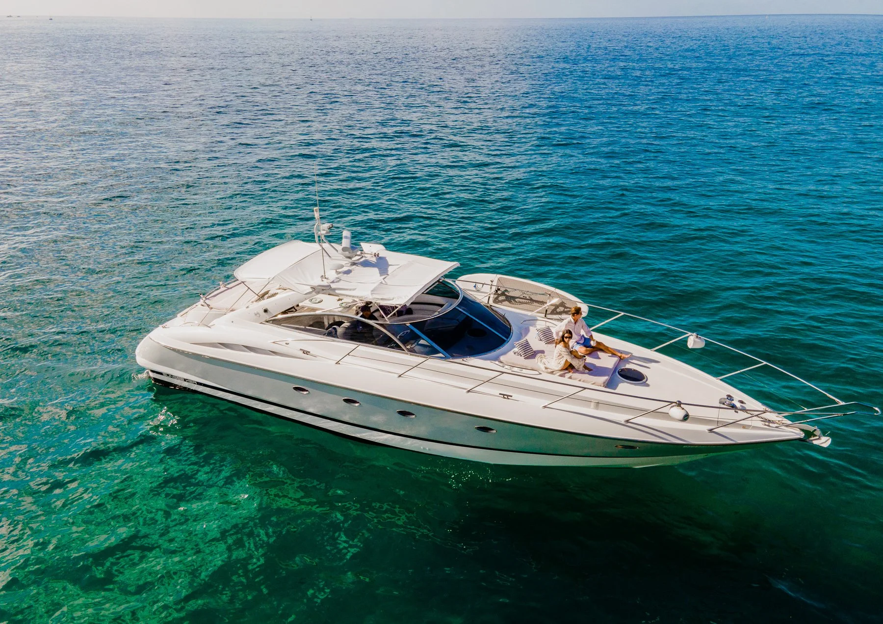 A white yacht with two people relaxing on the deck, floating on clear blue water under a bright sky.