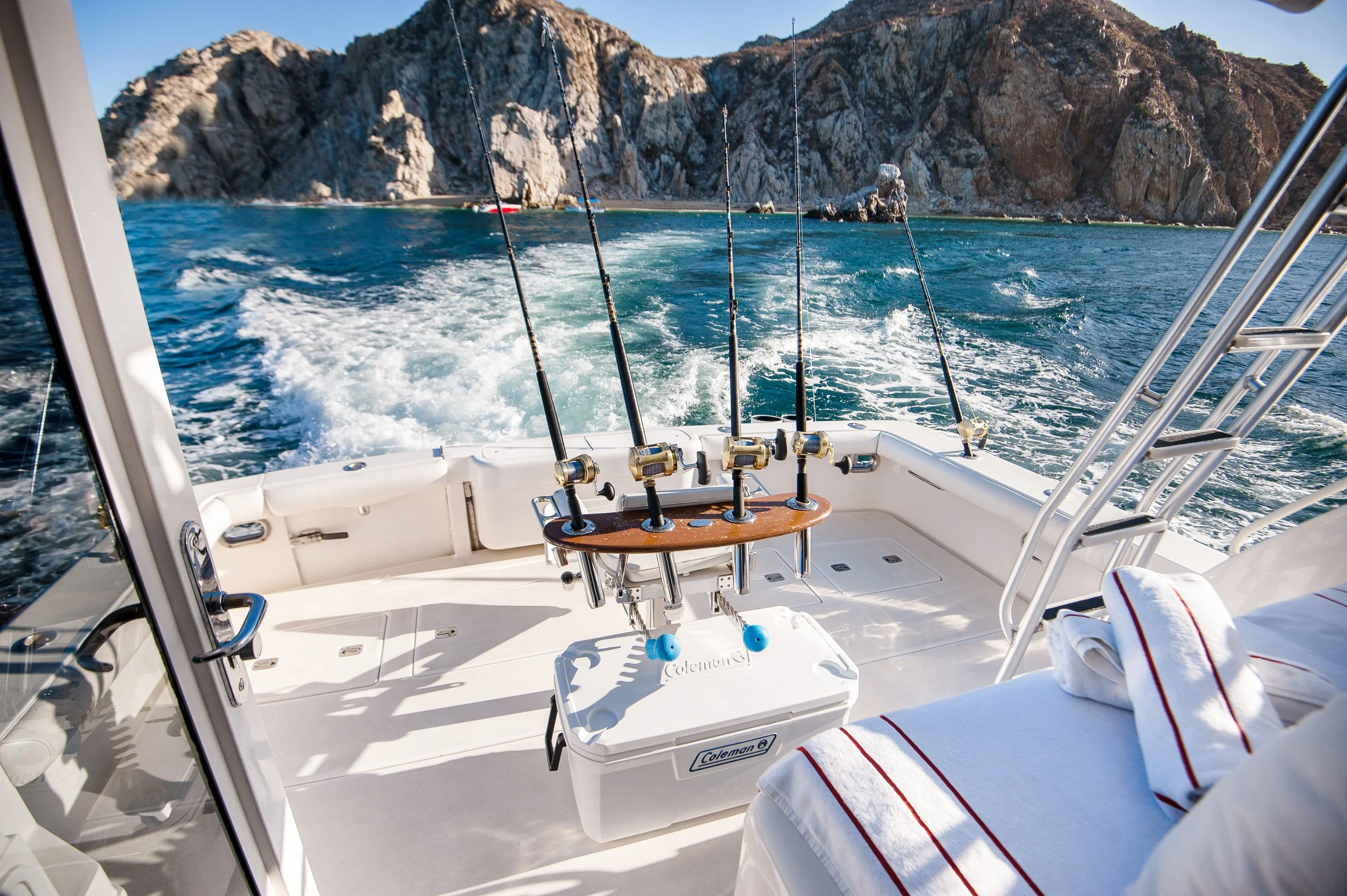 View from the back of a boat with four fishing rods secured in holders, overlooking blue water and rocky cliffs in the distance.