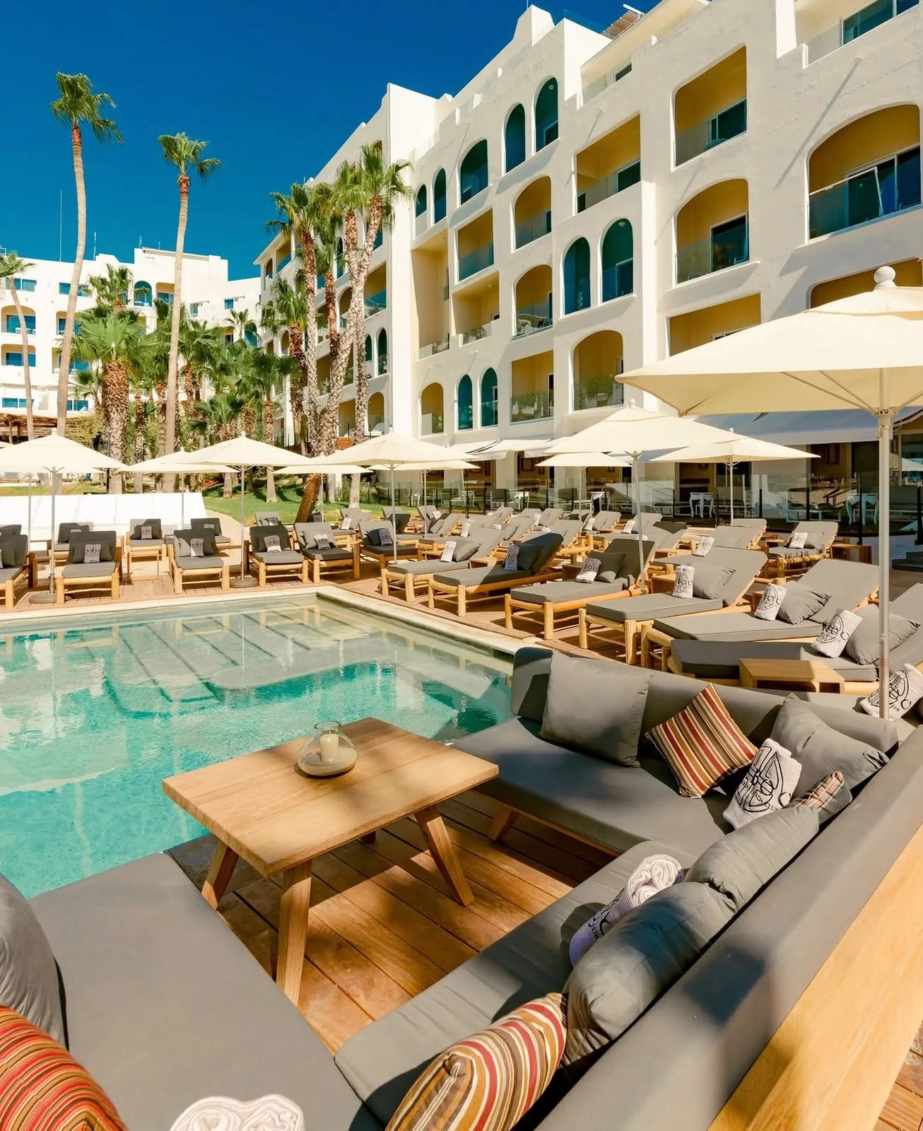 Luxury hotel poolside area with lounge chairs, umbrellas, and a building with white walls and green accents, surrounded by palm trees under a clear blue sky.