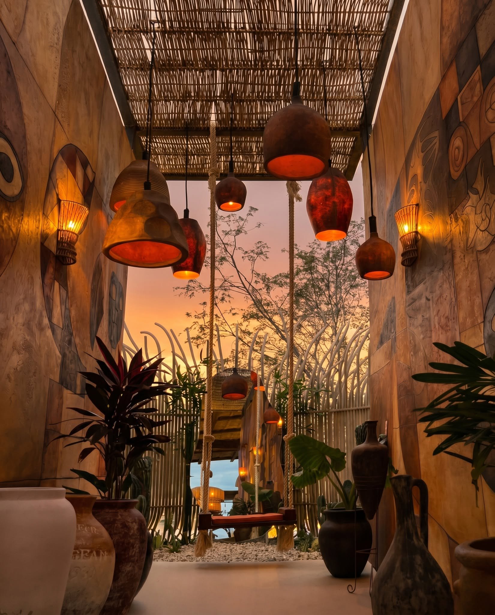 Interior view of a cozy space with hanging woven pendant lights, potted plants, and decorative vases, with a sunset visible through the window.