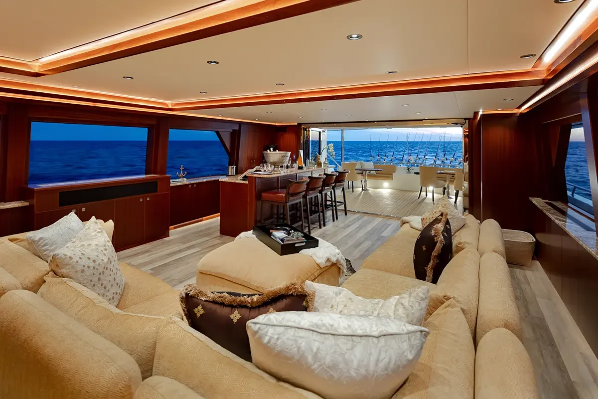 Luxury yacht interior with a large beige sectional sofa, throw pillows, a bar area, and ocean views through large windows.