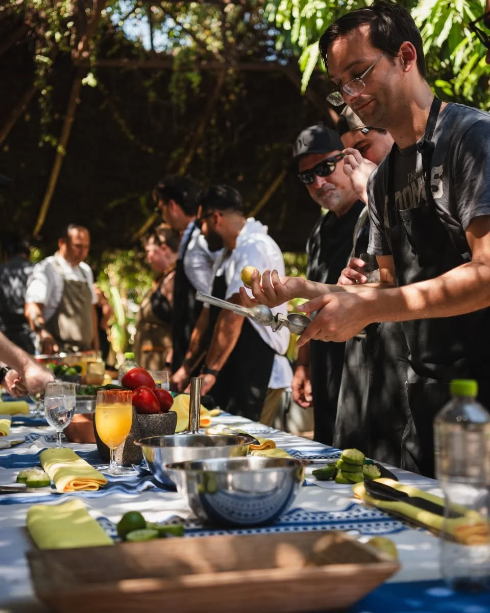 People gathered outdoors around a long table preparing food, with various bowls, fruits, and drinks on the table.