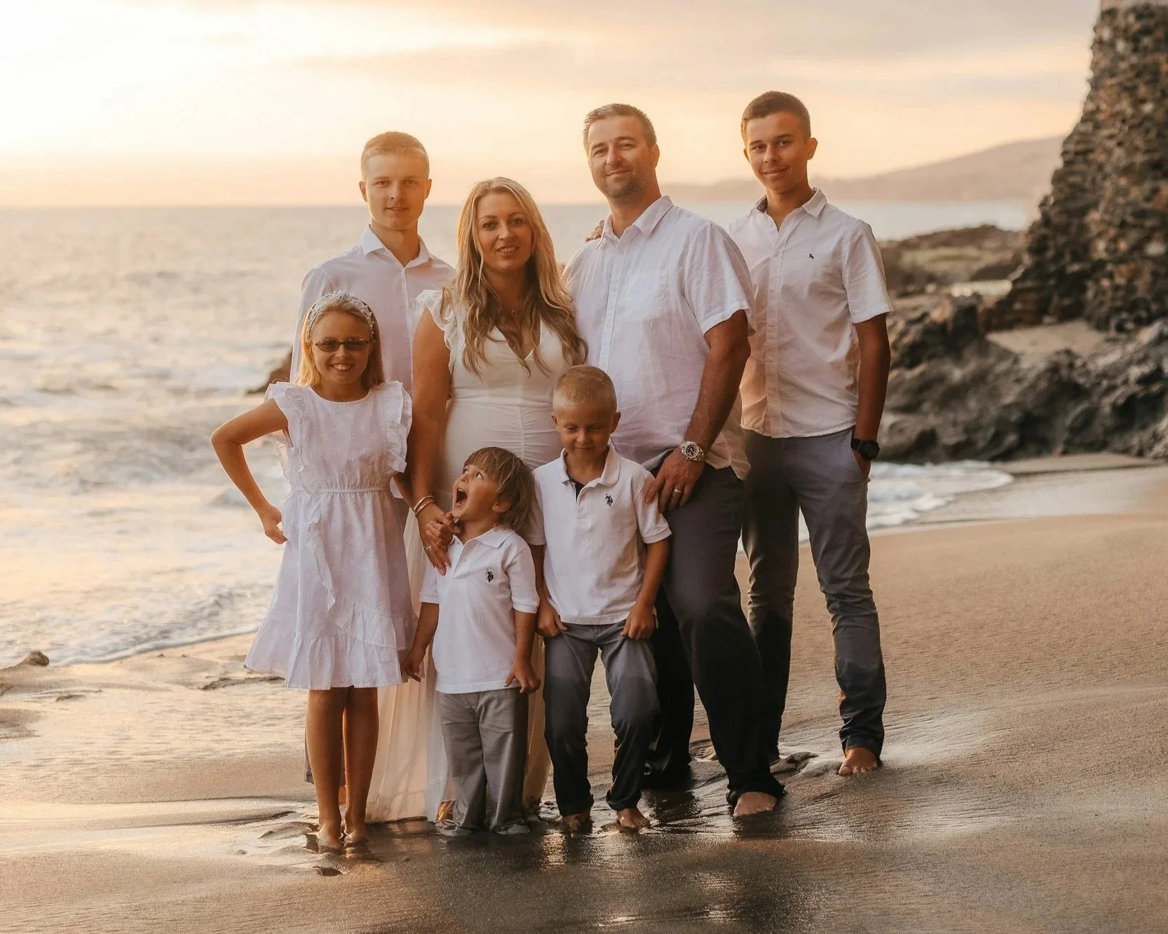 Family of nine standing on the beach at sunset, smiling and posing for a photo.