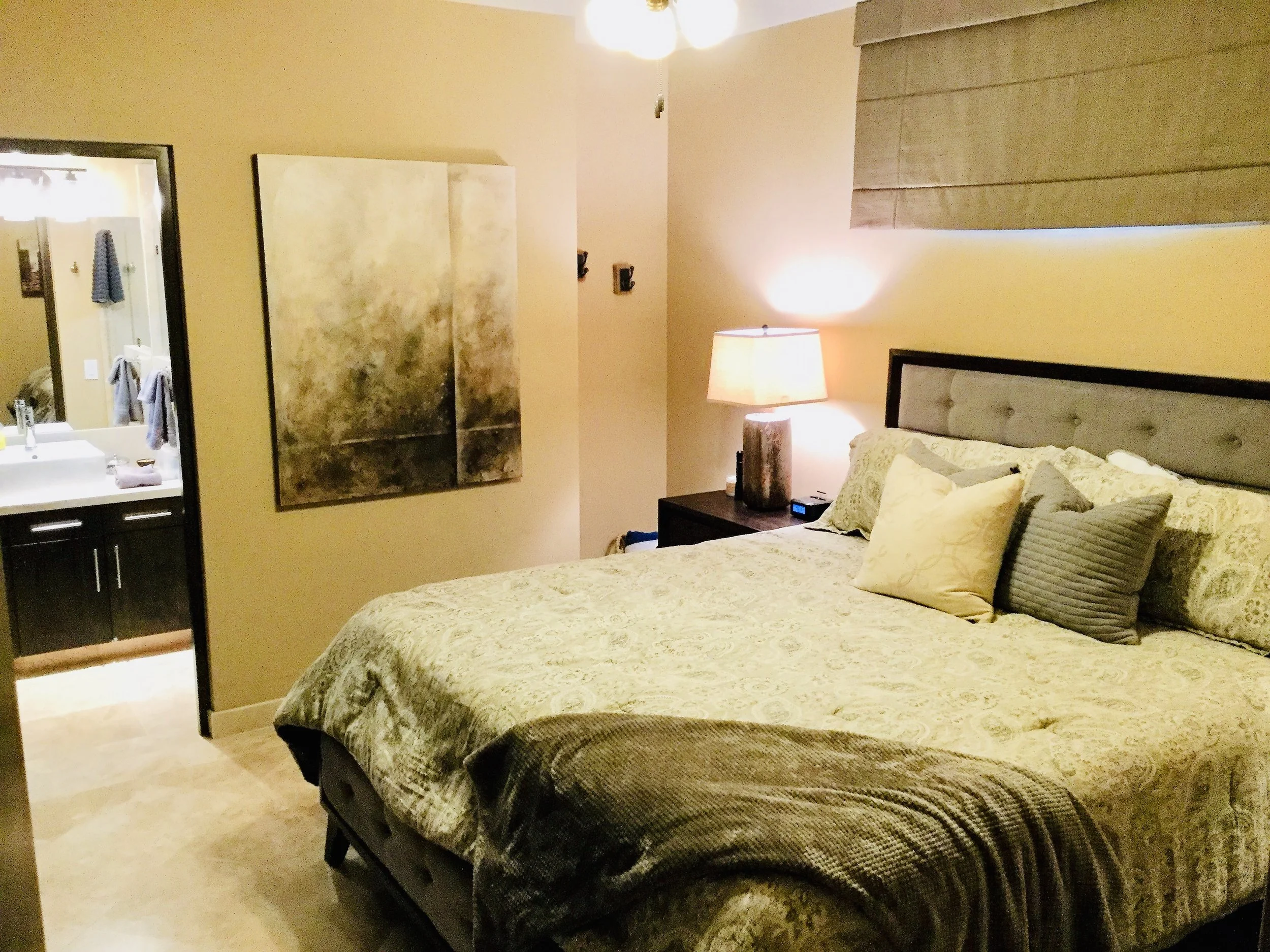 A bedroom with a bed covered with a patterned quilt and multiple pillows, a nightstand with a lamp, and a wall-mounted light. There's a large painting on the wall and a window with a pleated shade, with part of a bathroom visible through an open door.