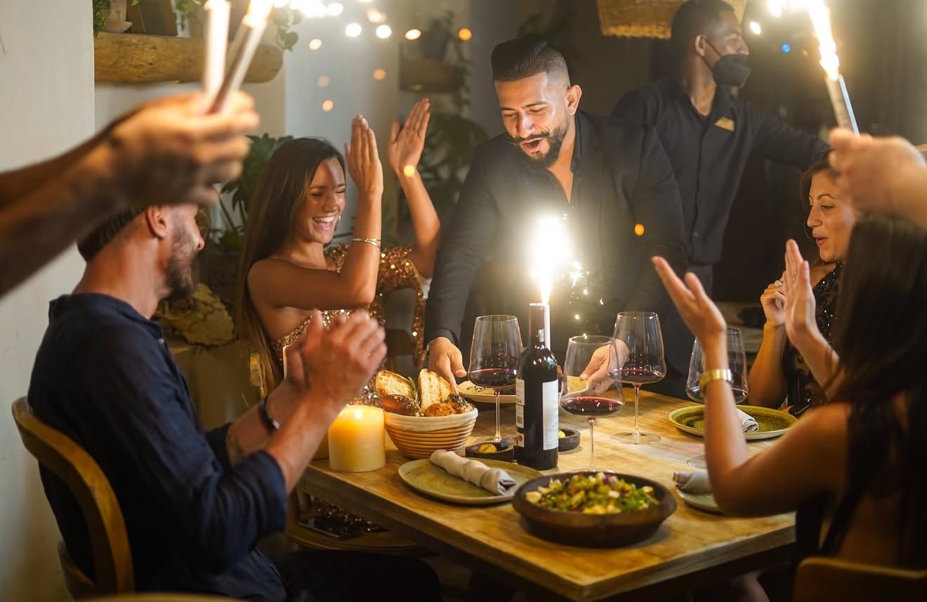A group of people celebrating a birthday with a cake, sparklers, and wine at a dinner table, smiling and cheering.