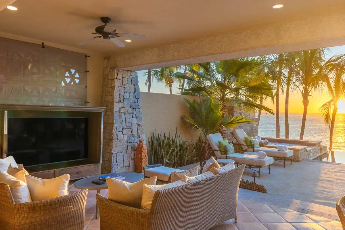 Outdoor living space with wicker furniture, a large TV, and lounge chairs by the ocean at sunset, surrounded by palm trees.