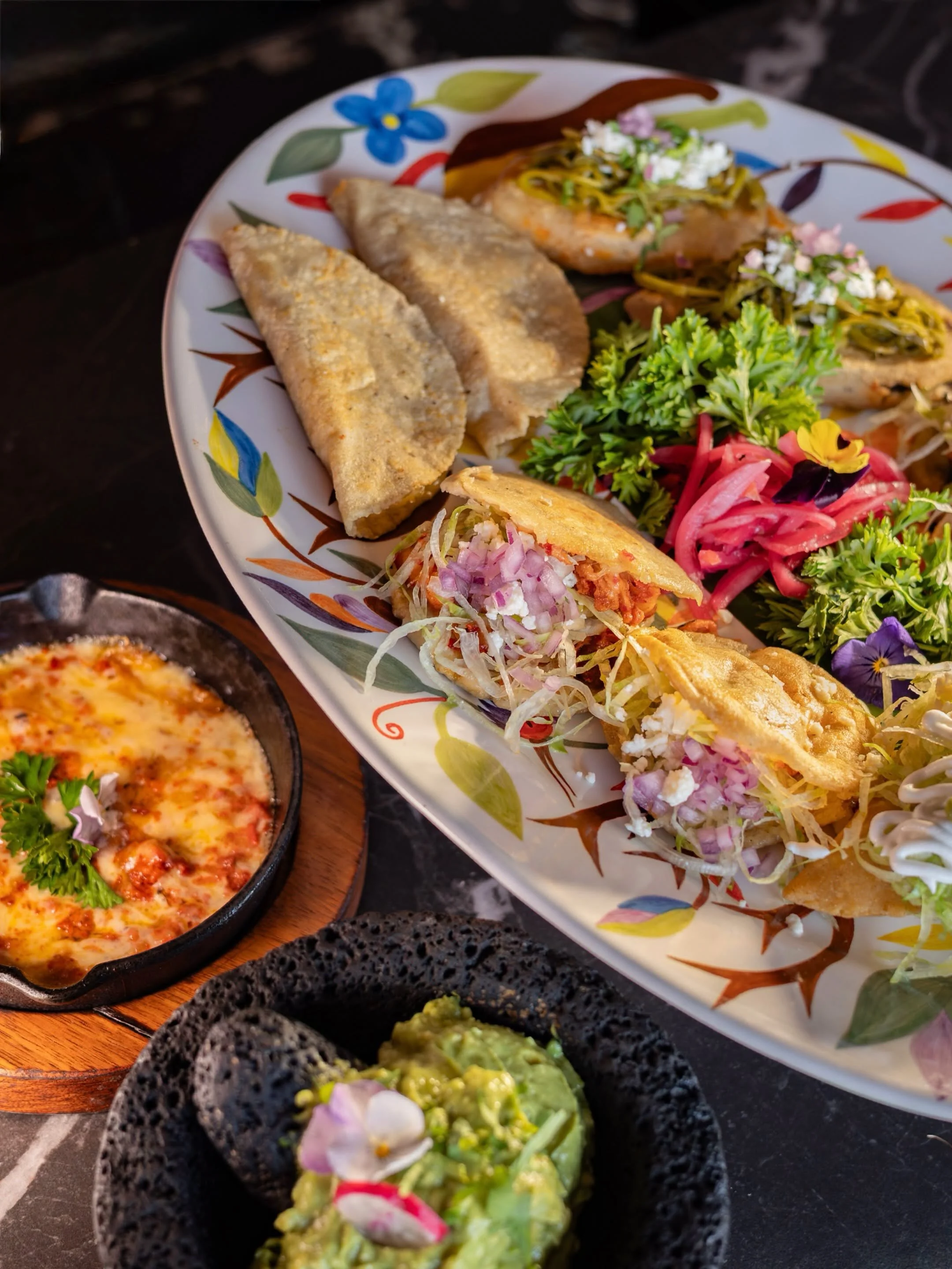 Plate of Mexican food with tacos, mini pizza, guacamole, and various garnishes on a dark surface.