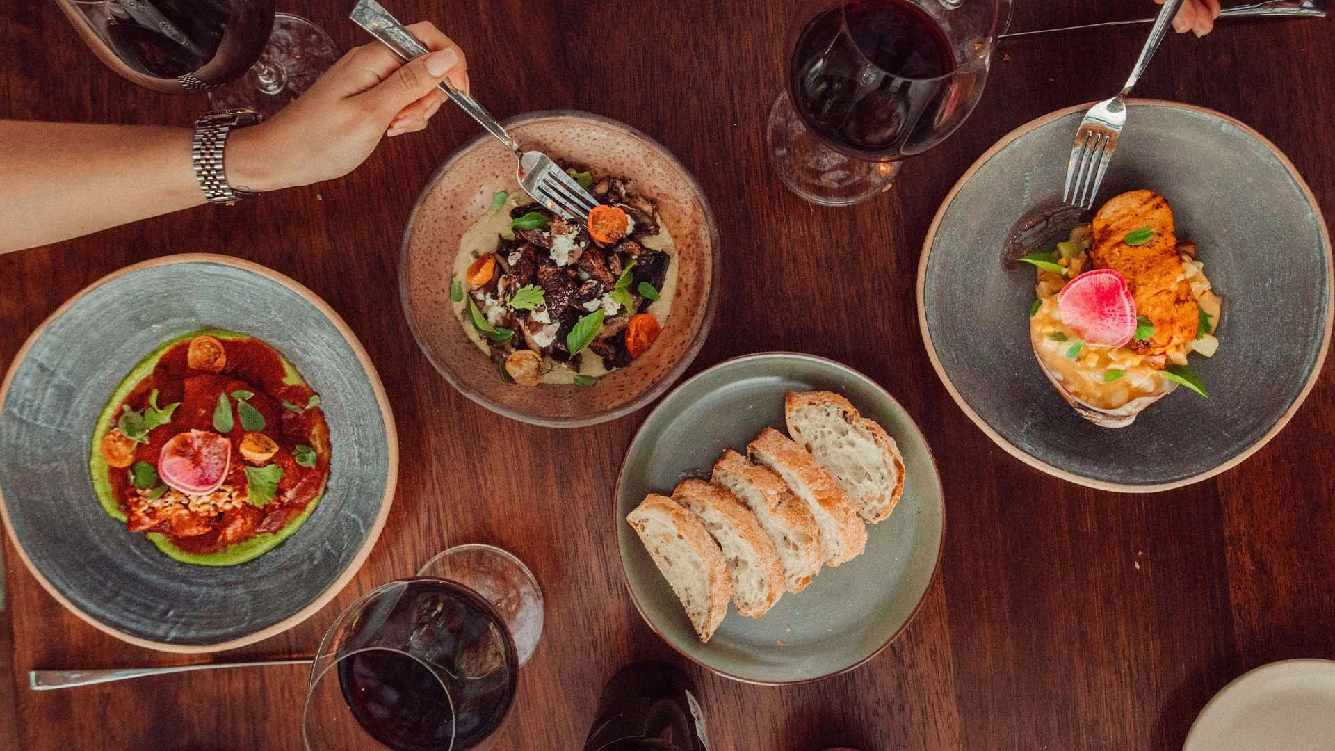 A table with various dishes and glasses of red wine. There are three main plates of food, including a salad, an appetizer with a roll of bread, and a plated main course with sauce, vegetables, and garnishes. A person's hand with a silver watch is see