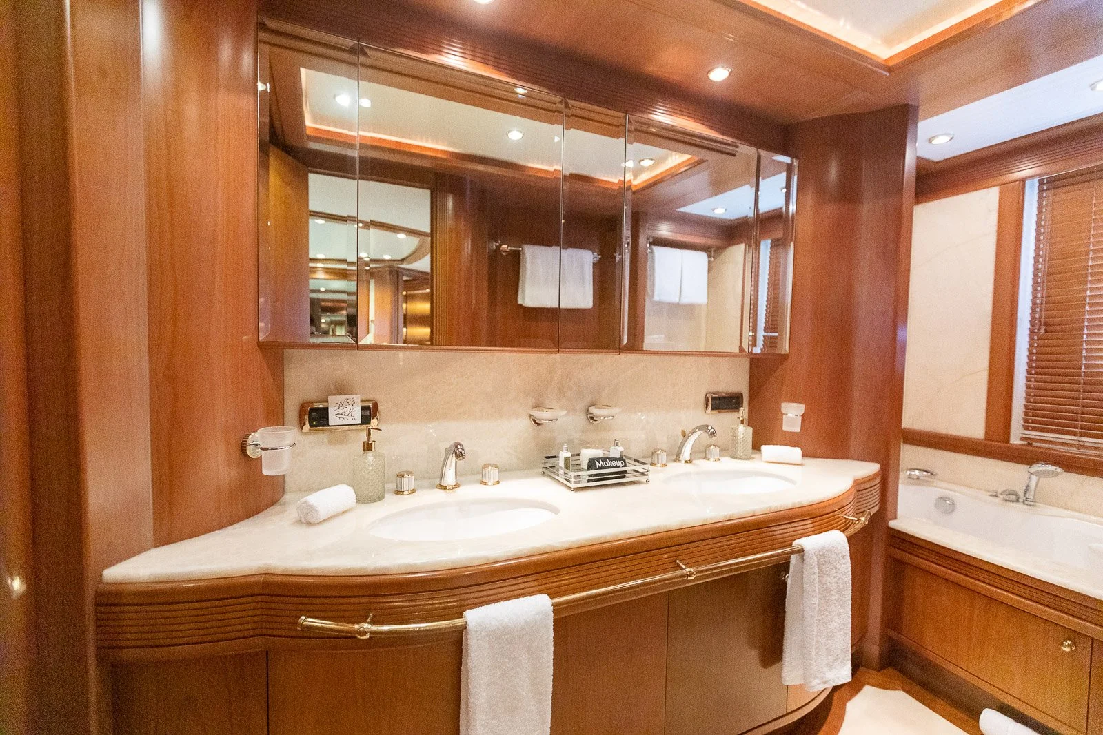 Luxurious bathroom with a double marble sink vanity, large mirror, and a built-in bathtub. Wooden cabinetry and trim, with natural light coming through blinds.