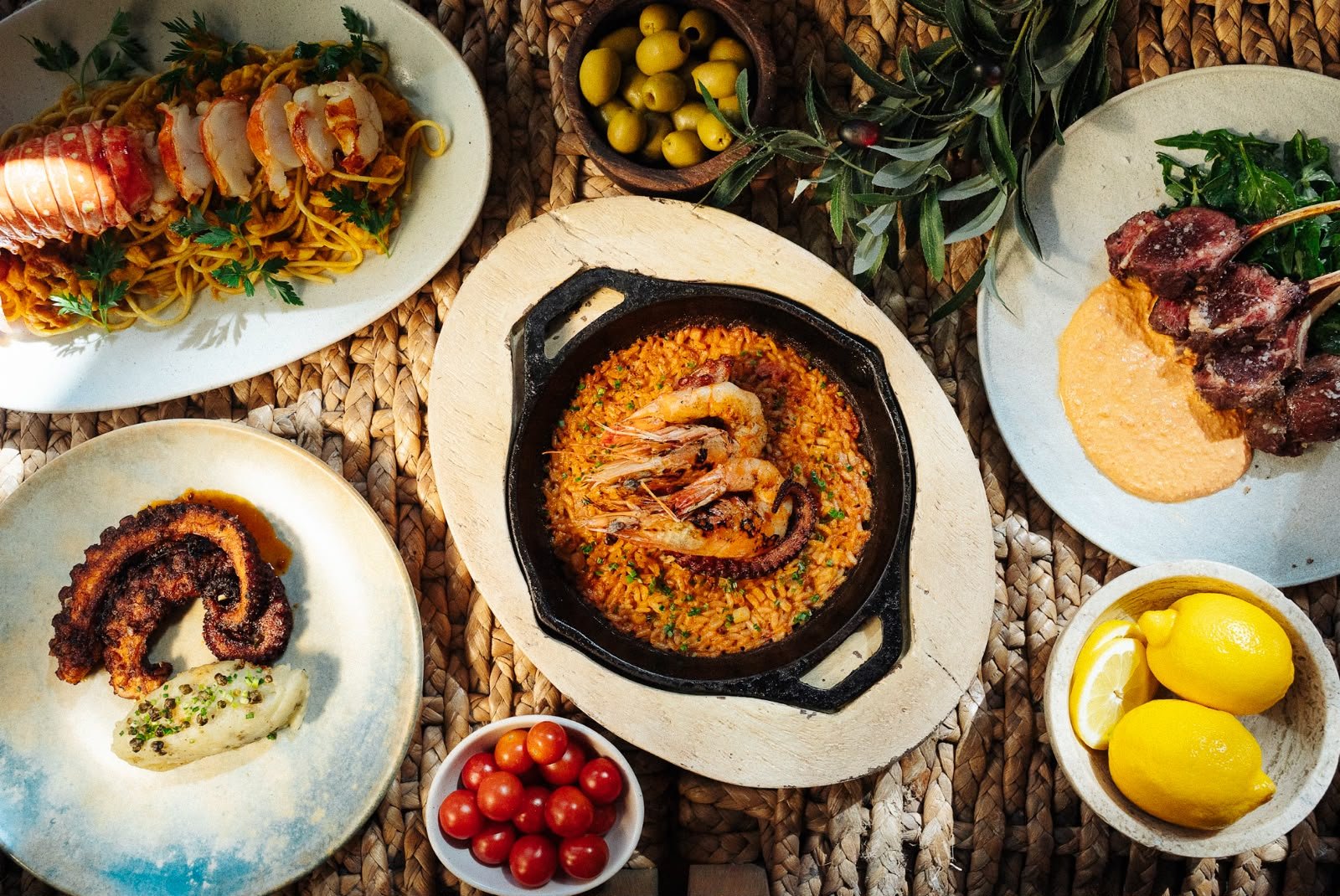 Assorted dishes including seafood paella, grilled octopus, lamb chops with sauce, and pasta with lobster, along with bowls of cherry tomatoes, lemons, olives, and leafy greens on a woven placemat.