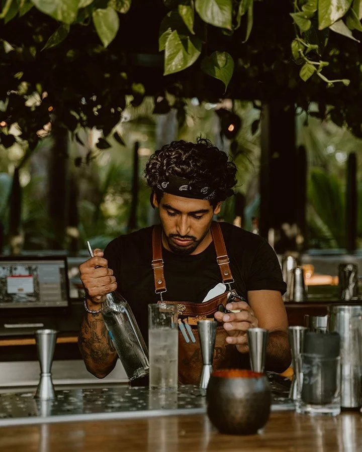 Bartender pouring a drink at an outdoor bar with a lush green canopy overhead.