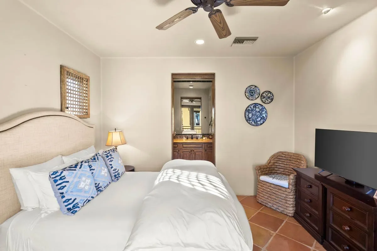 A bedroom with a large bed, blue and white pillows, a nightstand with a lamp, a wicker armchair, a flat-screen TV on a wooden dresser, and decorative plates on the wall.