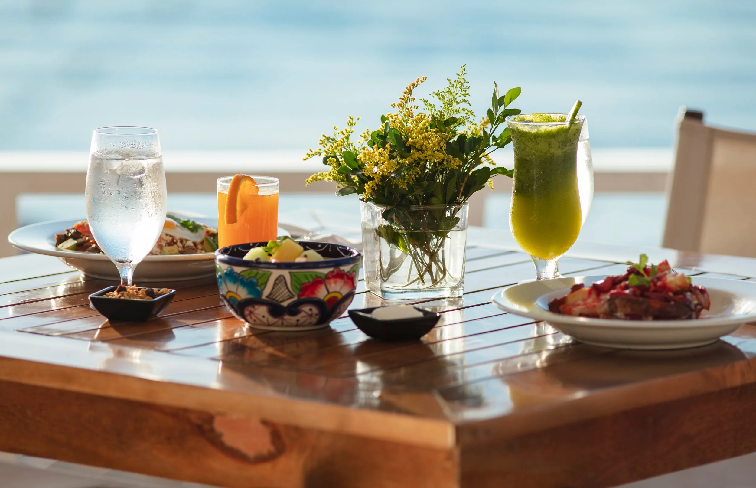 A wooden table set with various dishes including a glass of water, a glass of orange juice with an orange slice, a bowl of fruit, a glass of green smoothie, and plates of food, with a blue sky and ocean in the background.