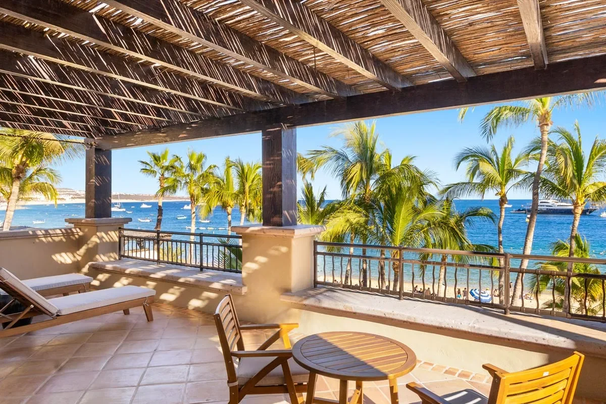 A tropical beach view from a shaded balcony with wooden furniture, including lounge chairs, a small table, and chairs, overlooking palm trees, the ocean, and boats.