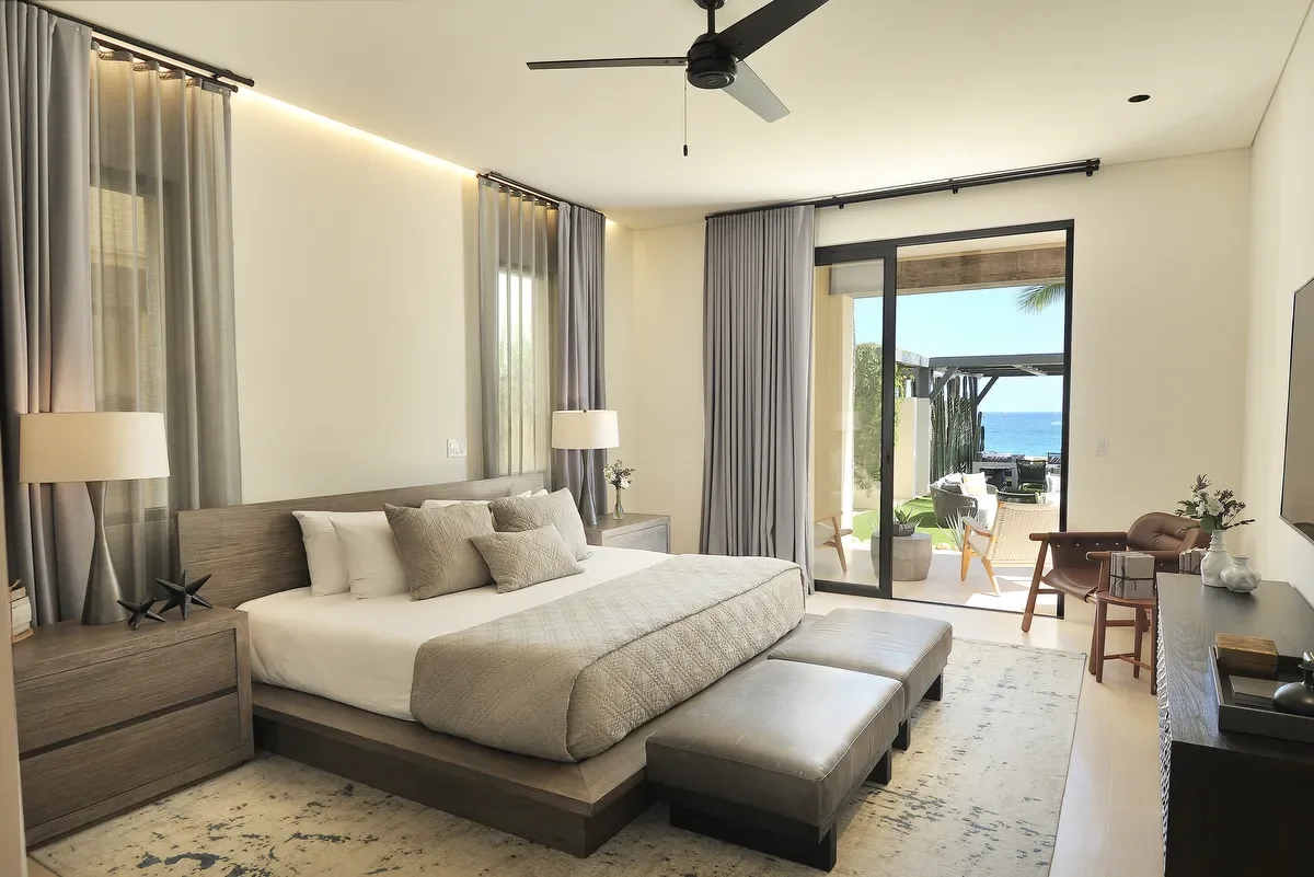 A modern bedroom with a large bed, nightstands, and lamps, opening to an outdoor patio with ocean view.