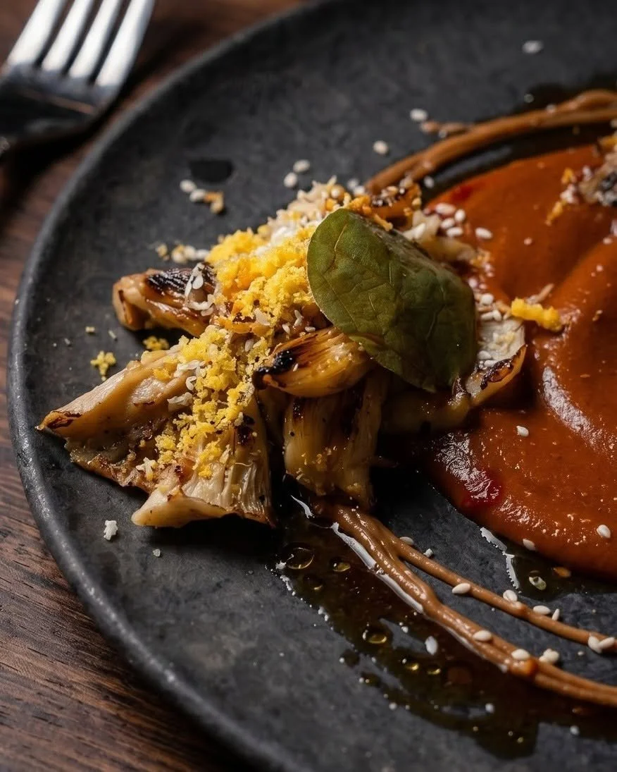 Close-up of a black plate with artichoke hearts, a bay leaf, sesame seeds, grilled vegetables, and a side of thick tomato sauce on a wooden table.