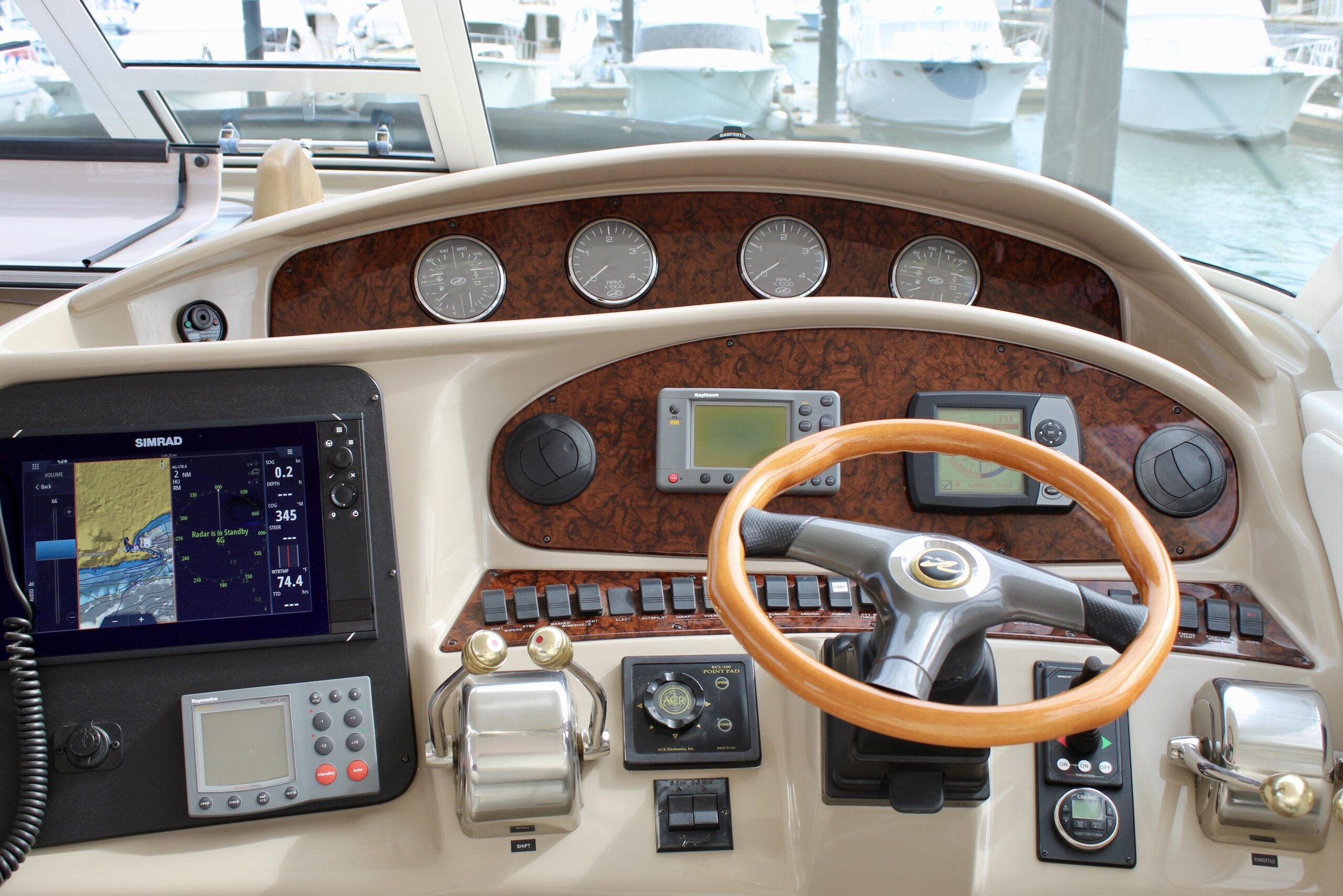 The image shows the helm of a boat with navigation and control instruments. There is a large wooden steering wheel, multiple gauges, electronic displays, and navigation devices mounted on a wood-paneled dashboard. In the background, through the winds