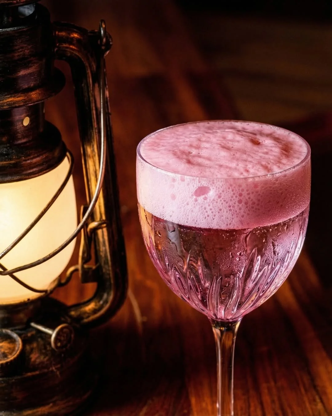 A pink cocktail in an elegant glass beside an antique lantern on a wooden surface.