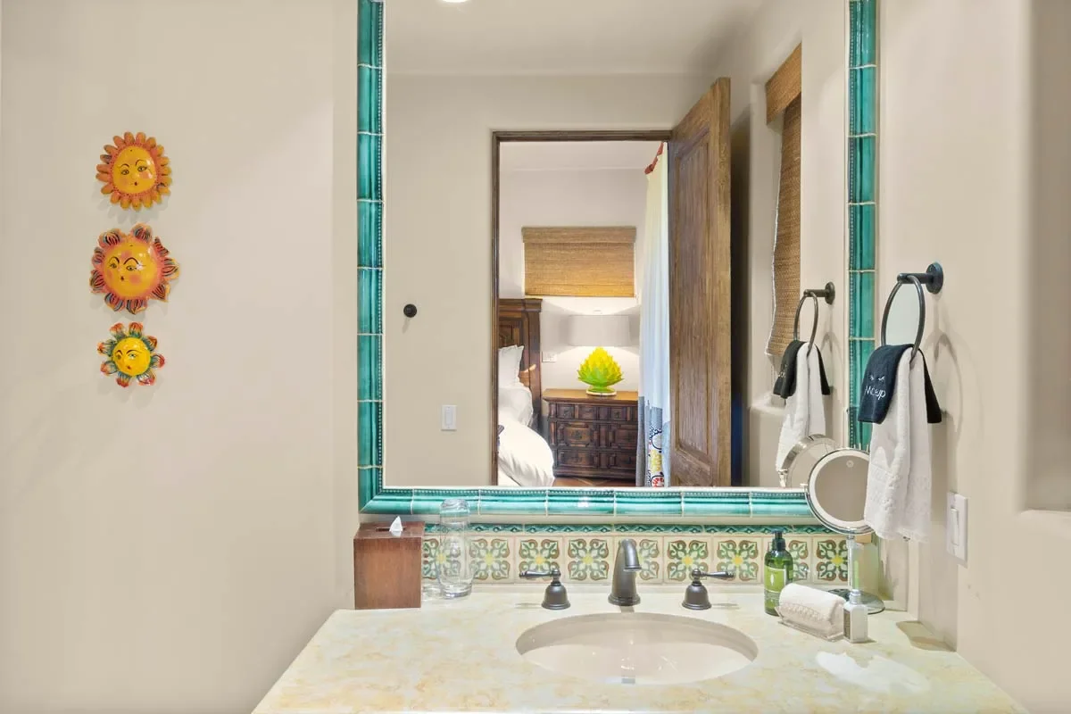 Bathroom vanity with beige countertop, dark fixtures, and a large mirror with decorative green and patterned tiles. Decor includes sun-shaped wall hangings and a glimpse of a bedroom with a wooden headboard and a yellow decorative piece.