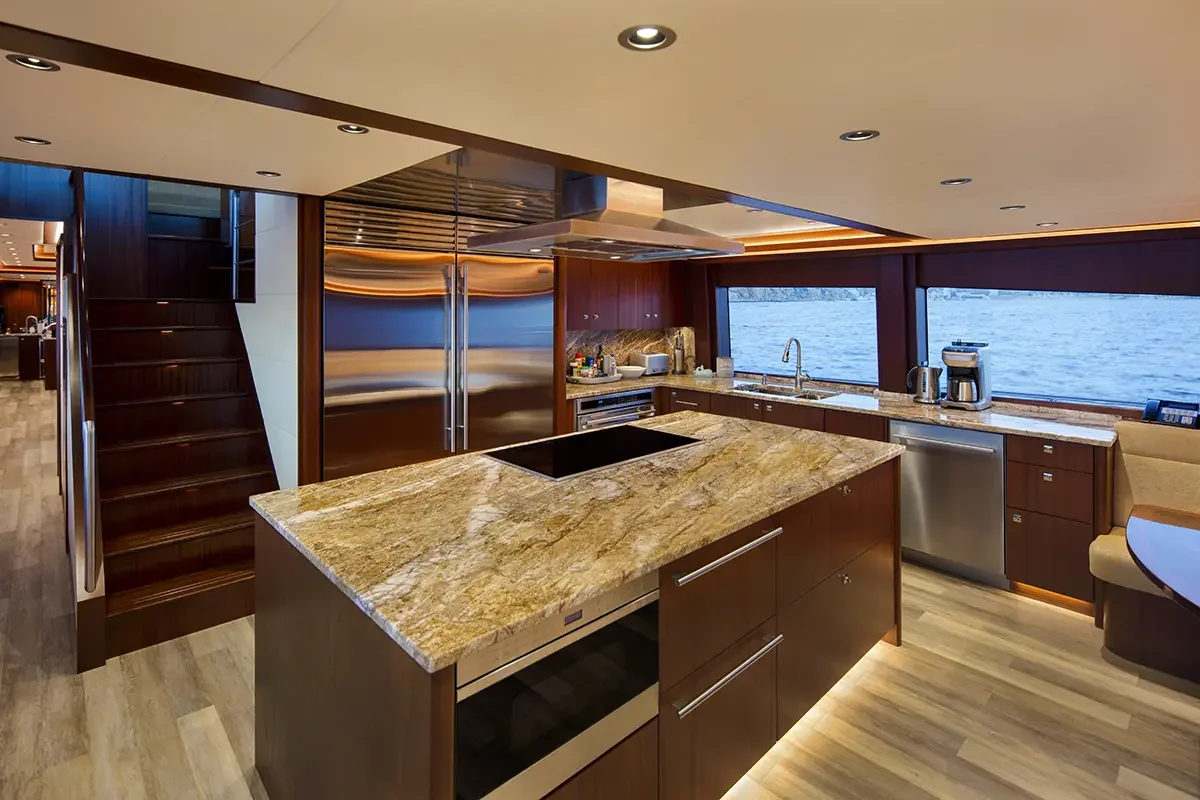 Modern kitchen with wooden cabinetry, a marble countertop island, stainless steel appliances, and ocean view through large windows.