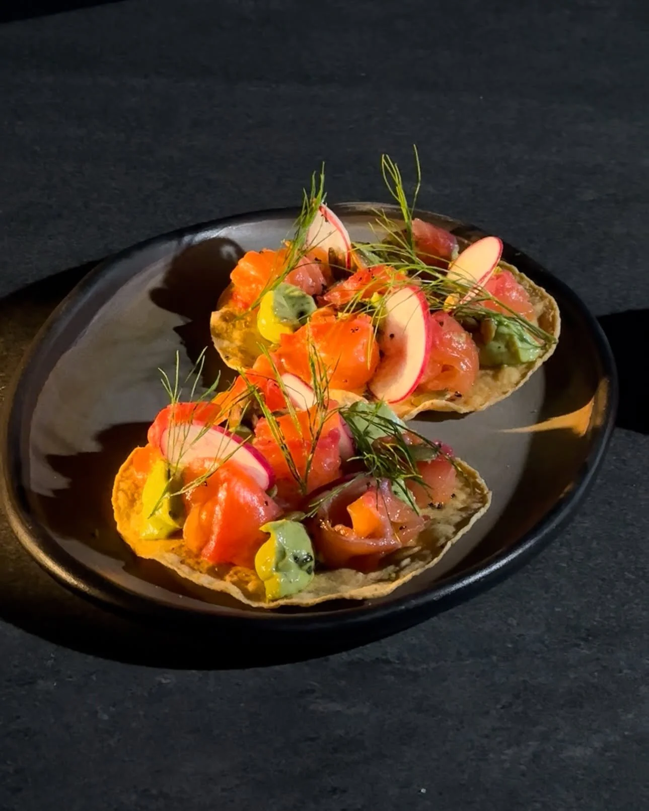 Tacos topped with slices of radish, salmon, avocado, and herbs on a black plate