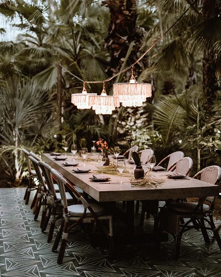 An outdoor dining table set for a meal with wine glasses, black plates, and decorative items, illuminated by a hanging fringe chandelier, surrounded by lush green tropical plants.