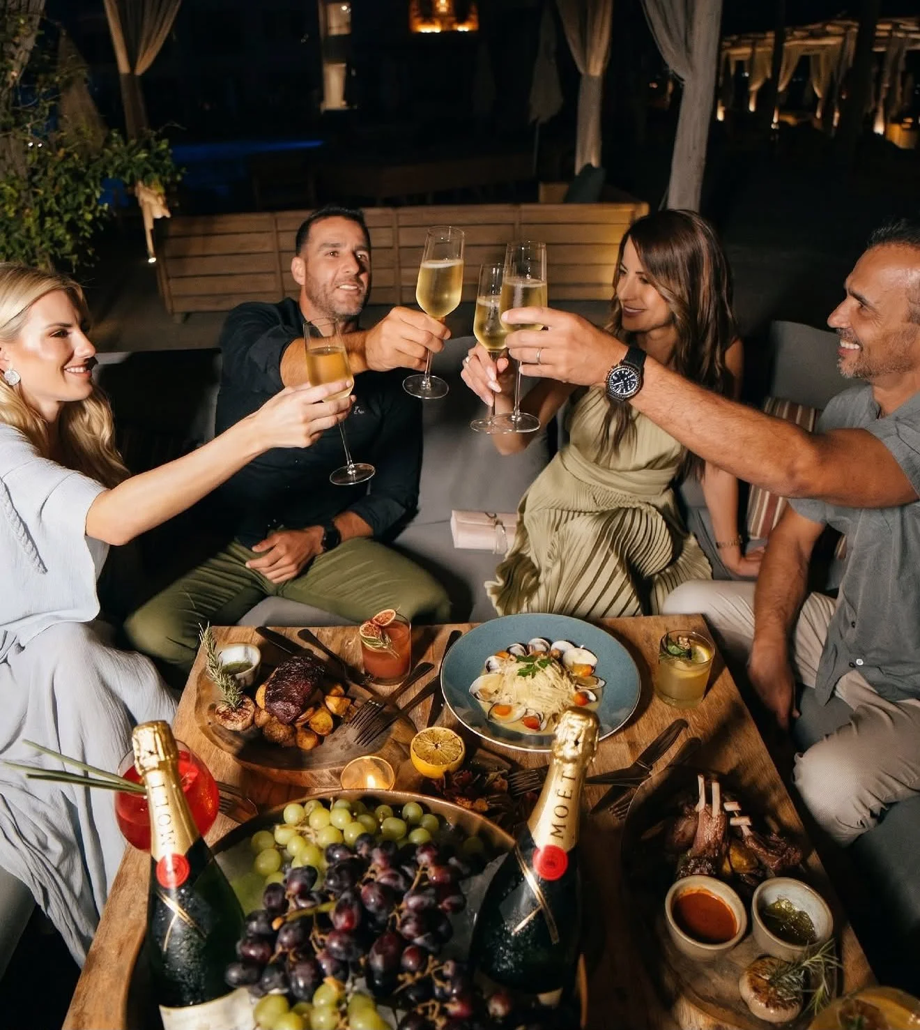 Group of five friends enjoying a dinner and raising champagne glasses in a toast at a cozy evening gathering with food and drinks on the table.