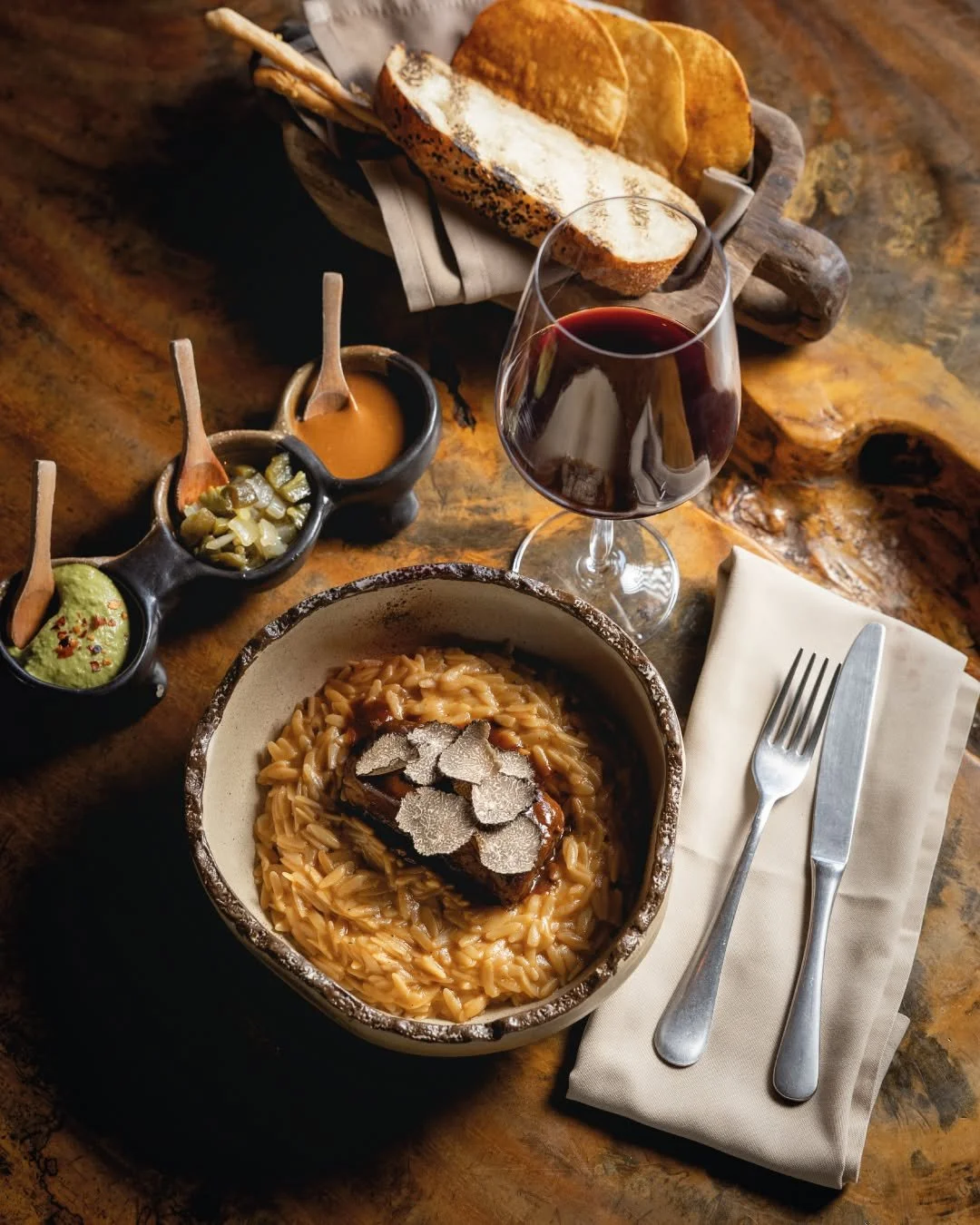 A bowl of pasta topped with truffle slices, served with a glass of red wine, bread, and small dishes of sauces and guacamole on a rustic wooden table.