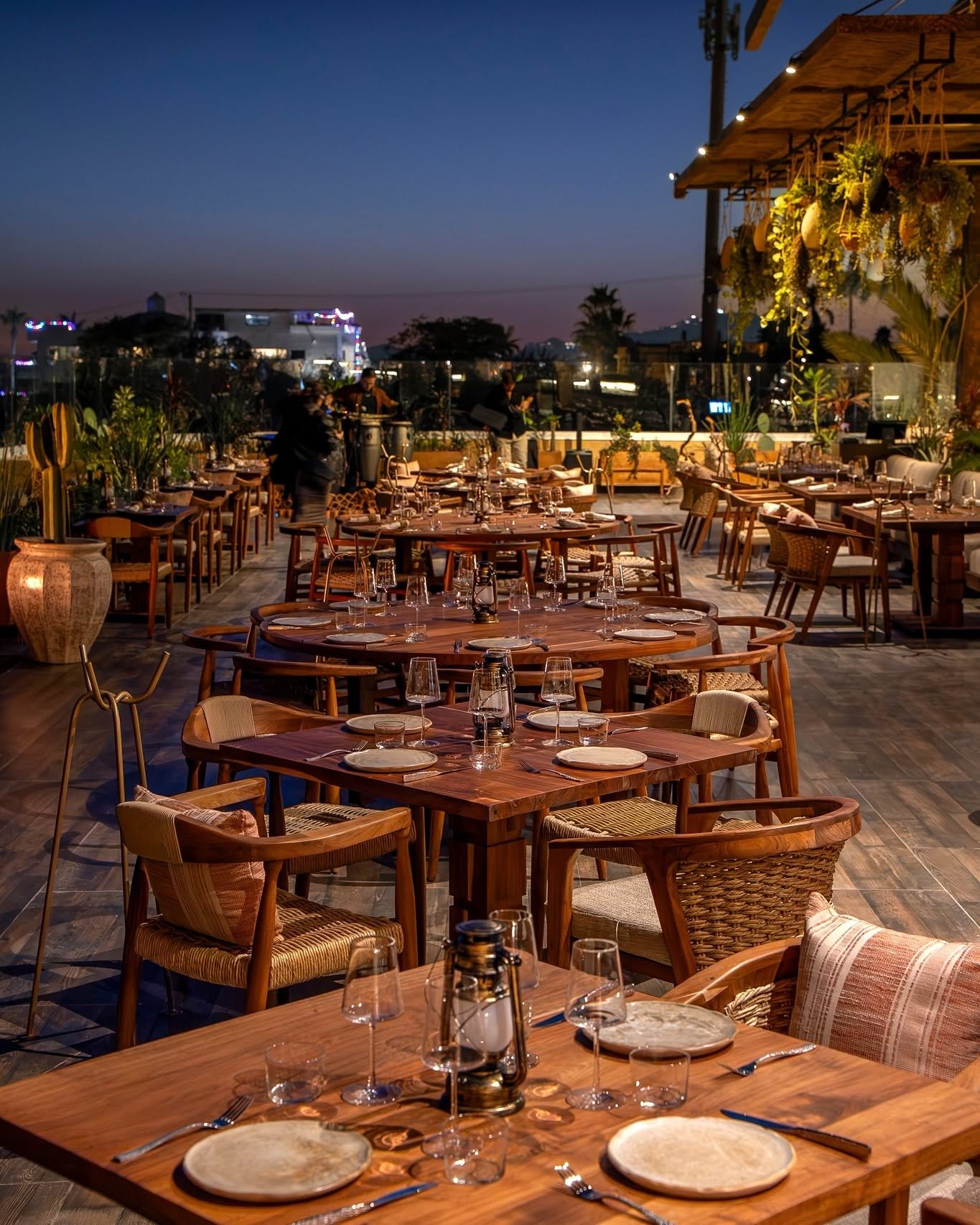 An outdoor restaurant terrace at dusk with elegantly set tables, wooden chairs, indoor plants, and string lights in the background.