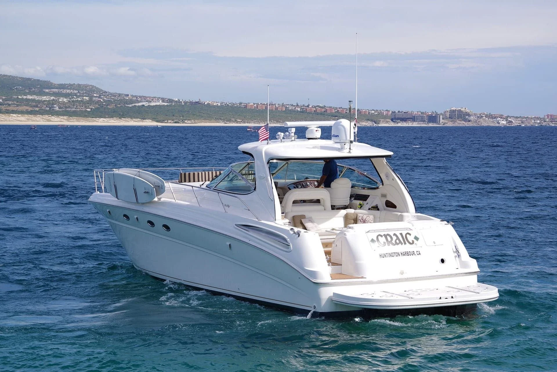 A white luxury motor yacht named CRAIC sailing in Huntington Harbour, California, with a green hilly coastline and buildings in the background.