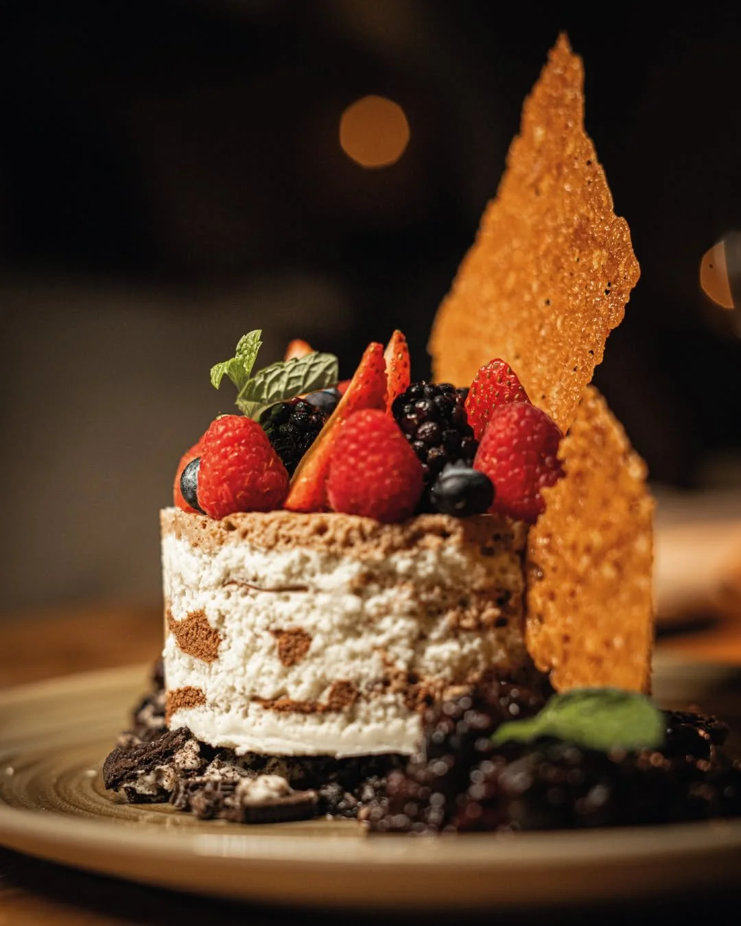 A dessert cake topped with strawberries, blackberries, and fresh mint leaves, decorated with wafer cookies and chocolate shavings on a golden plate.