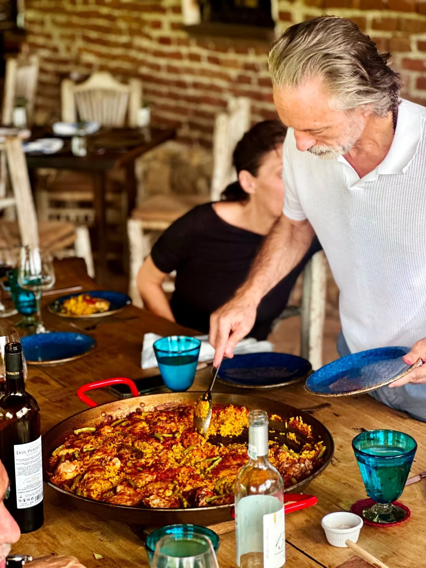 A group of people at a dinner table with a large pan of paella, various drinks, and plates, in a rustic restaurant setting with a brick wall.