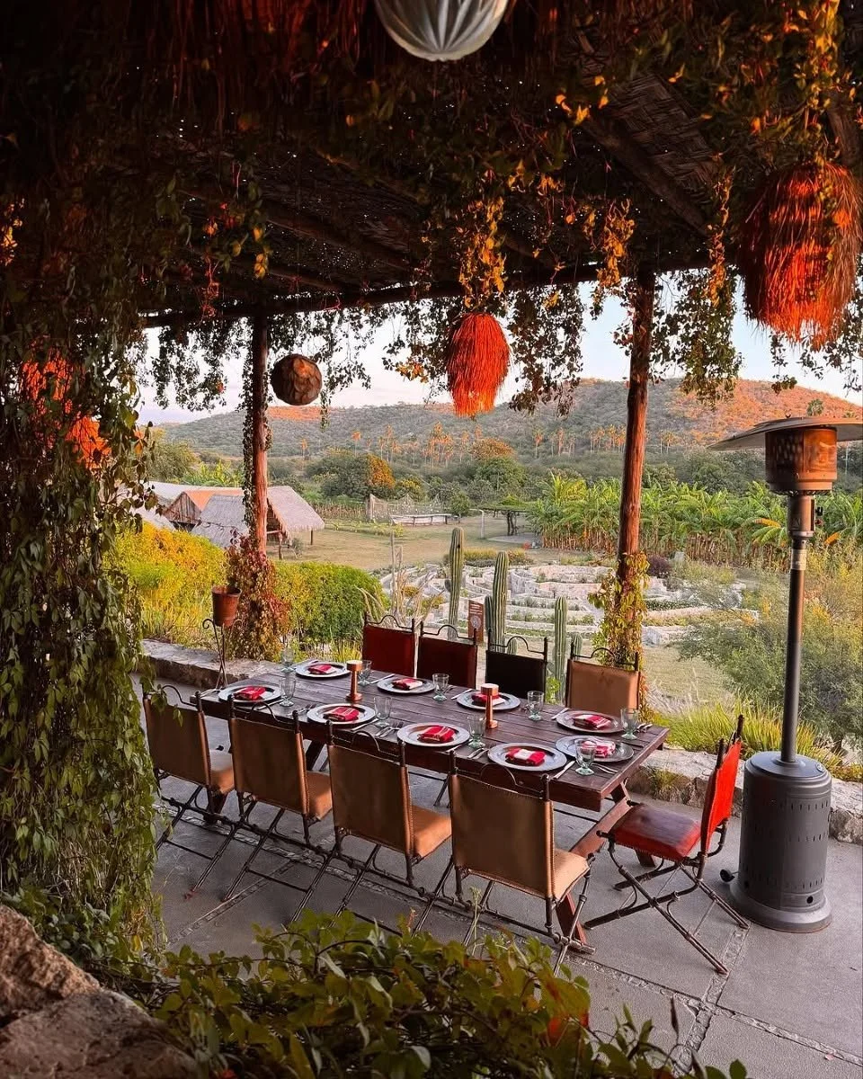 Outdoor dining setup with a long wooden table and nine chairs, set with plates, glasses, and red napkins, under a thatched roof with hanging decorations. Green landscape and hills visible in the background.