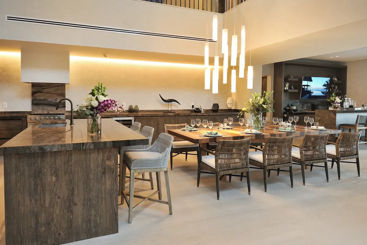 Modern dining room with a large wooden table set with glassware and floral centerpieces, surrounded by woven wicker chairs, and a kitchen island with chairs in a stylish home interior.