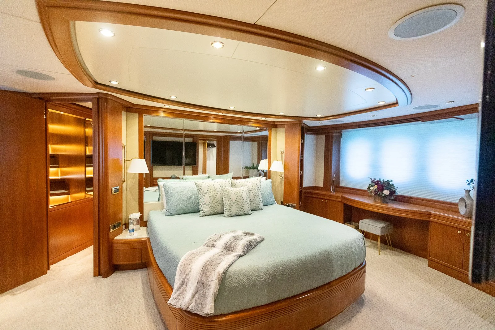 Luxury bedroom with a large bed, wooden paneling, mirrored closet doors, wall lamps, a flat-screen TV, a window with a blind, and decorative flower arrangements.