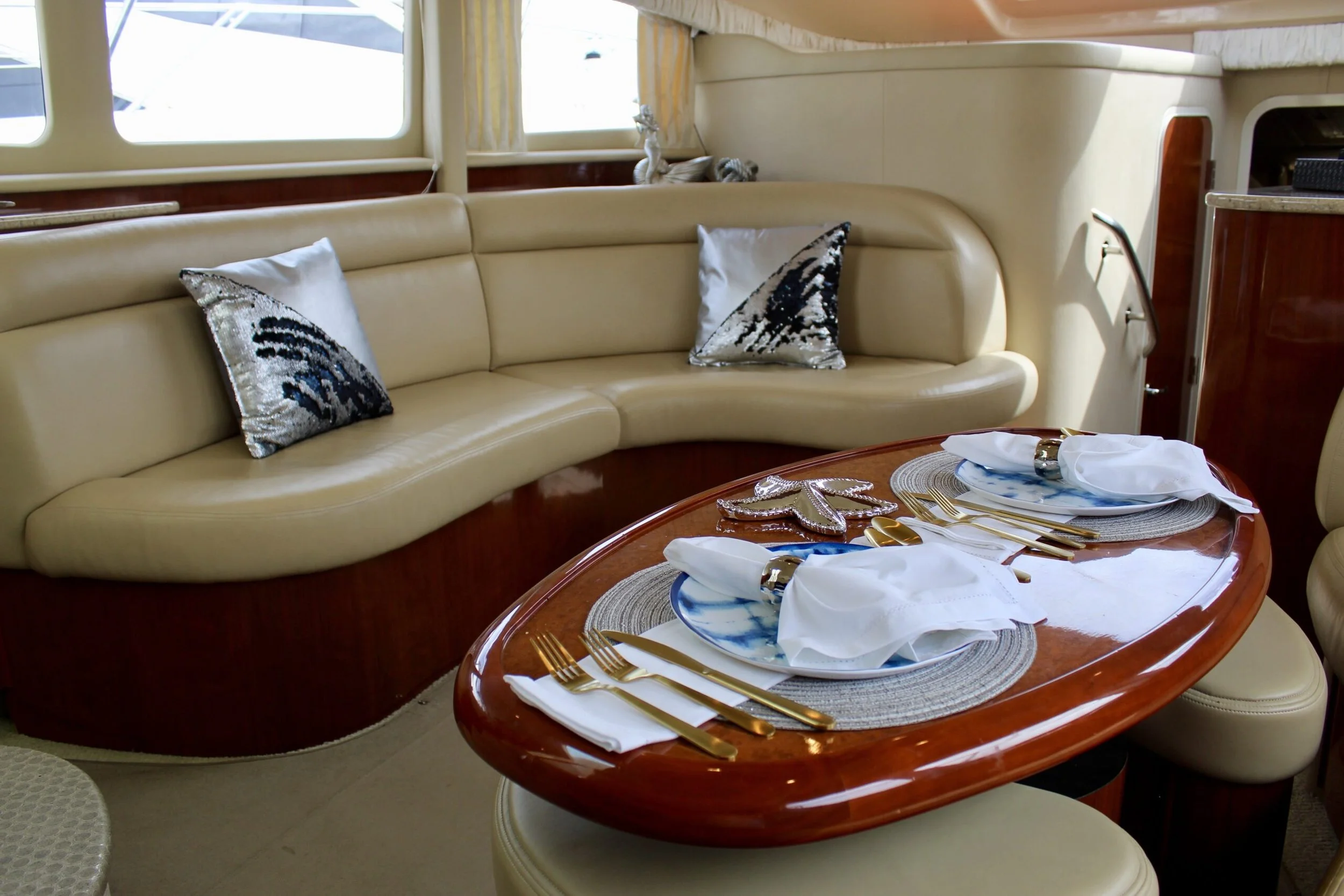 A beige curved sofa with decorative pillows, positioned in a room with large windows. In front, a polished wooden dining table is set with white napkins, gold utensils, and blue and white plates, suggesting a dining setup in a yacht or boat interior.
