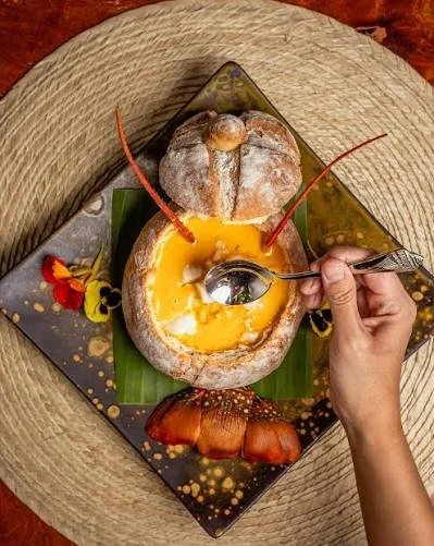 A person using a spoon to scoop custard from a hollowed-out baked pastry, decorated with cherries and powdered sugar, on a decorative black and gold plate with floral accents, placed on a wooden table.