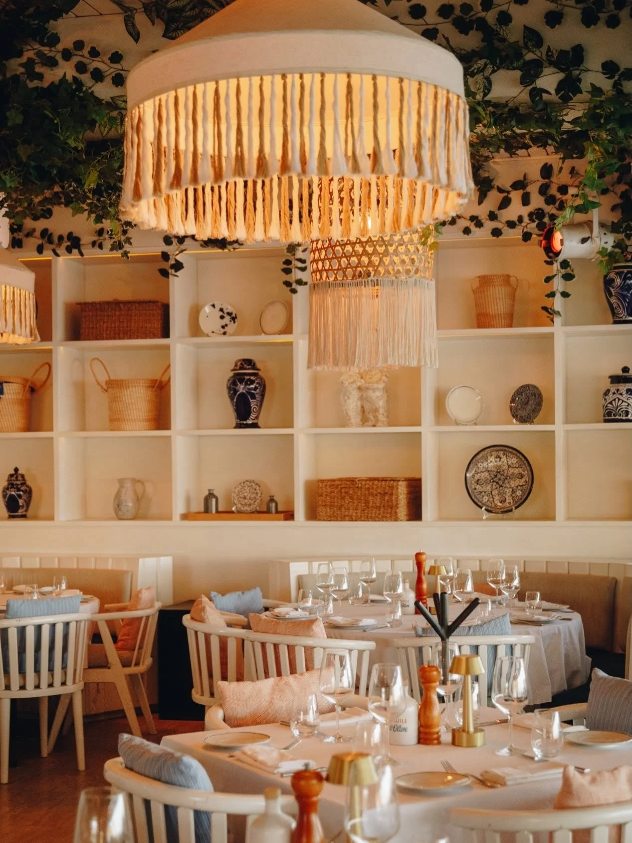Elegant restaurant interior with round tables set with glassware and utensils, surrounded by white wooden chairs with cushions. Decor includes shelves with baskets, vases, and decorative plates, and a large ceiling lamp with a fabric fringe and tassels, along with hanging green plants.