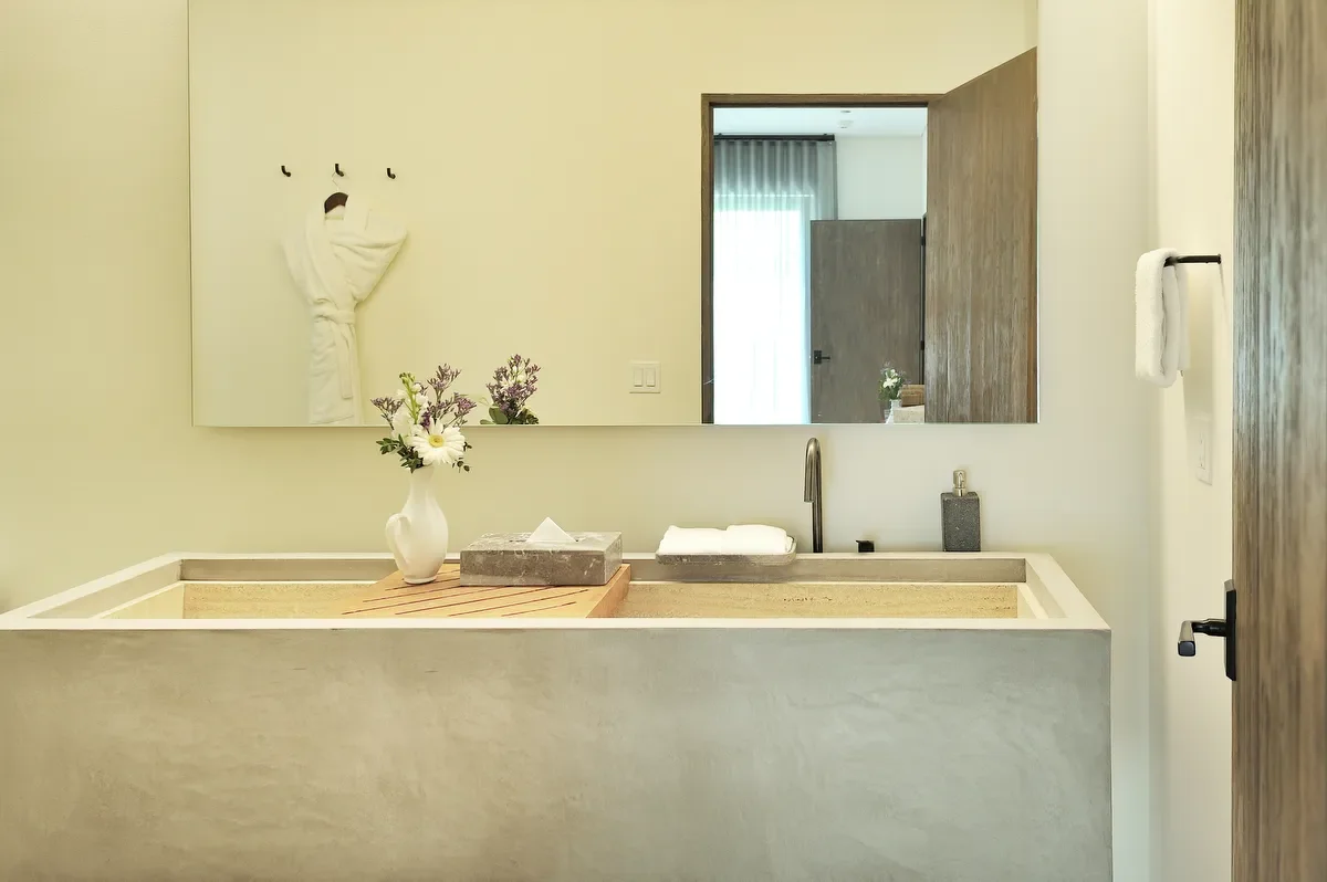 Bathroom with a large mirror above a white sink, a vase with flowers, a soap dispenser, and a towel on a black towel bar, with a doorway leading to a room with a closed curtain.