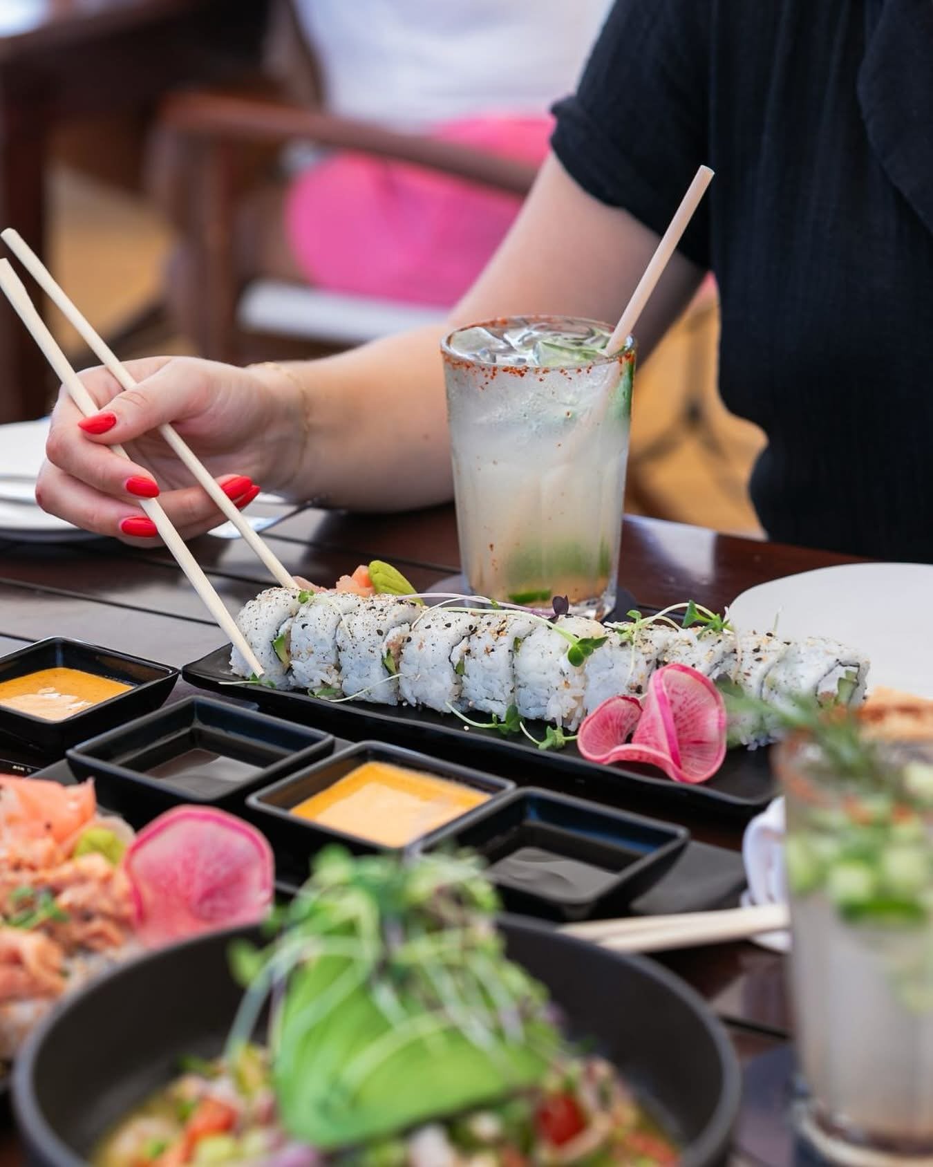 A person with red-painted nails is using chopsticks to pick up sushi rolls from a table of Japanese dishes, including soy sauce, and a glass of cocktail with lime and herbs.
